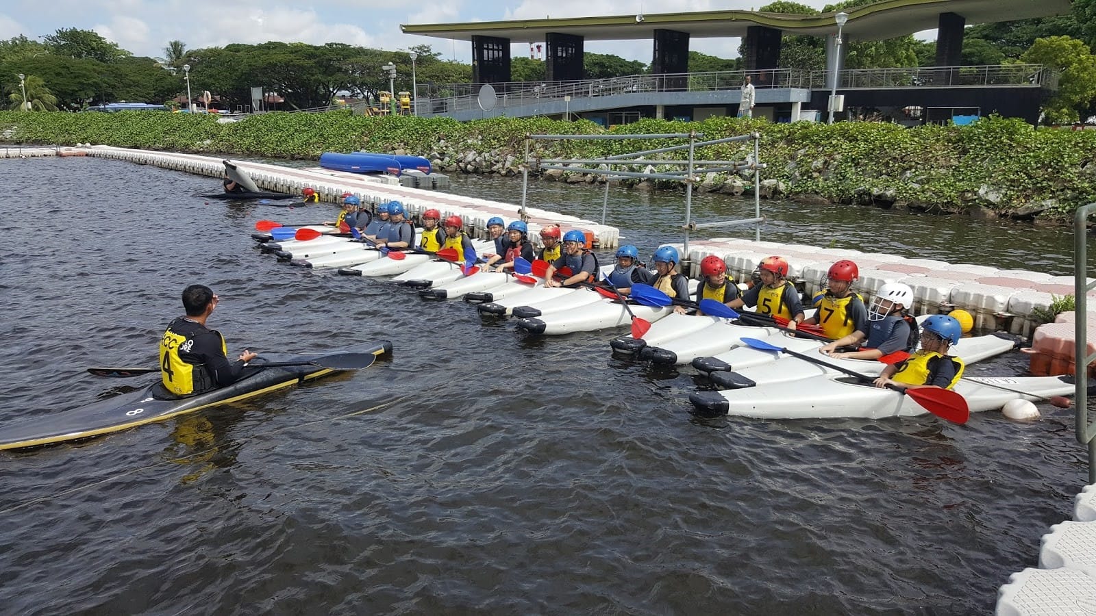 A group of students sits in individual kayaks lined up along a floating dock in a calm water area. Each student wears a life jacket and safety helmet, holding a paddle across the kayak. An instructor in a kayak faces the group, appearing to give guidance or instructions.